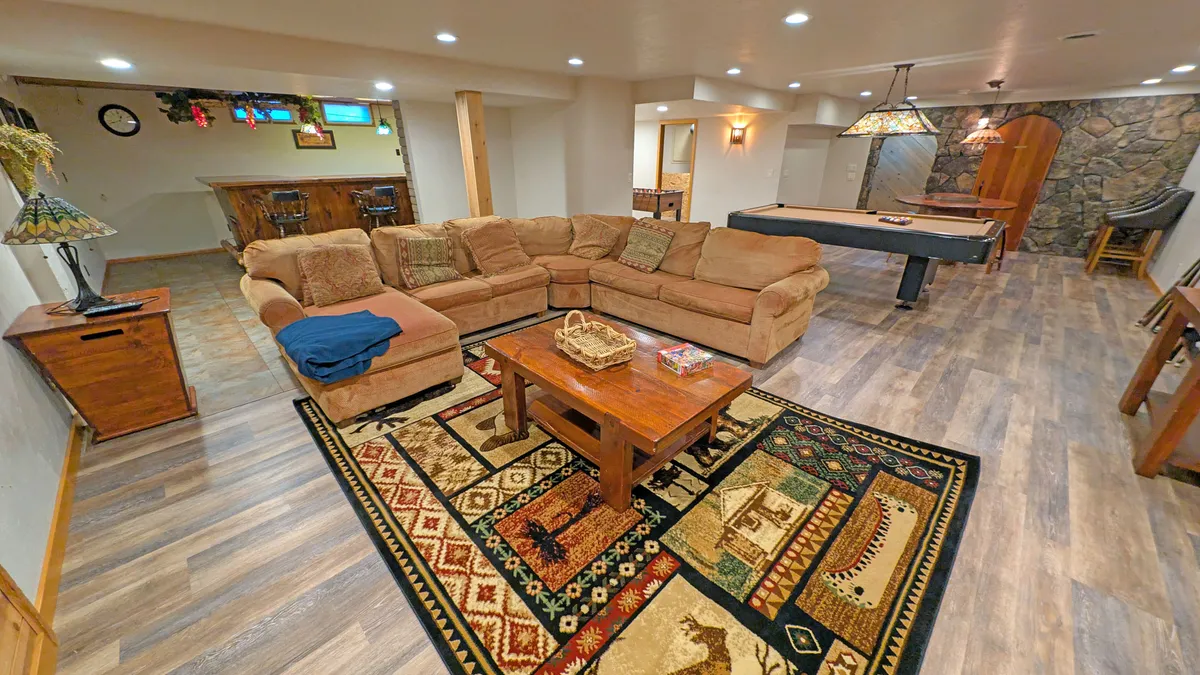 This image captures the heart of the entertainment basement: a large sectional sofa arranged around a wooden coffee table, with a colorful area rug adding vibrancy. The pool table and a cozy bar area are visible in the background, indicating a multi-functional space designed for both relaxation and fun. The overall atmosphere is comfortable and social, perfect for movie nights, group games, or celebrations.