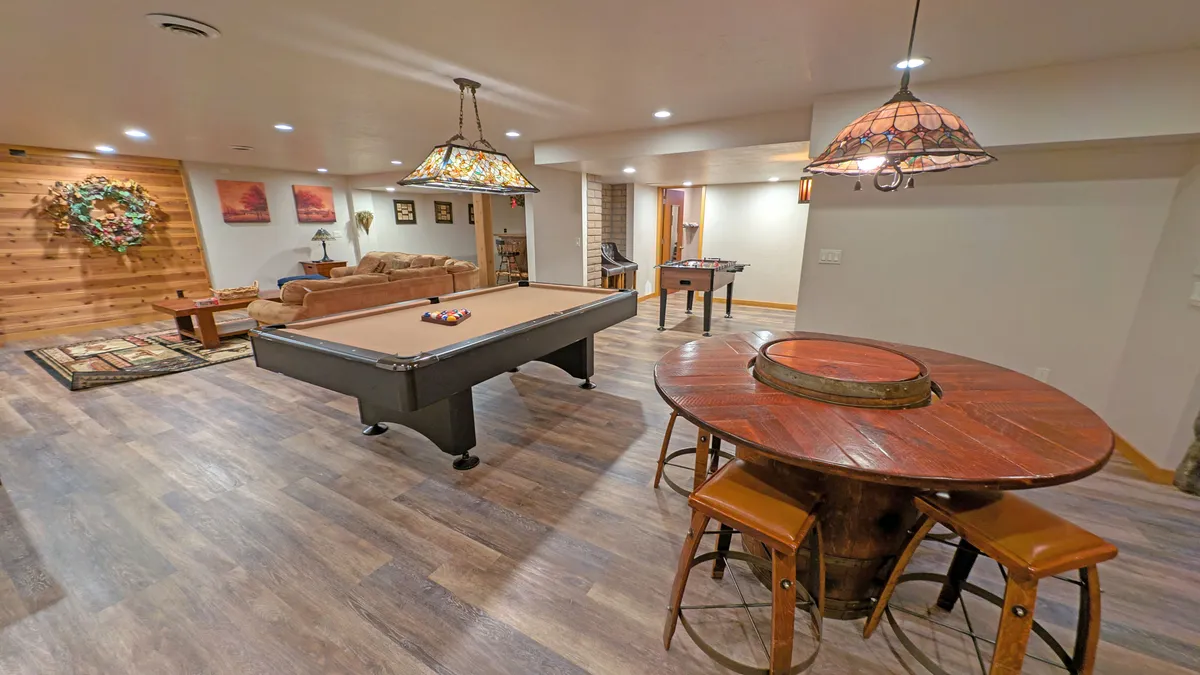 The image showcases an inviting, open-concept living space with a pool table as the centerpiece, plush couches for lounging, a large dining table perfect for group meals, stylish wall art, and warm hanging lights. The setting radiates a casual, social atmosphere ideal for gatherings and family fun.