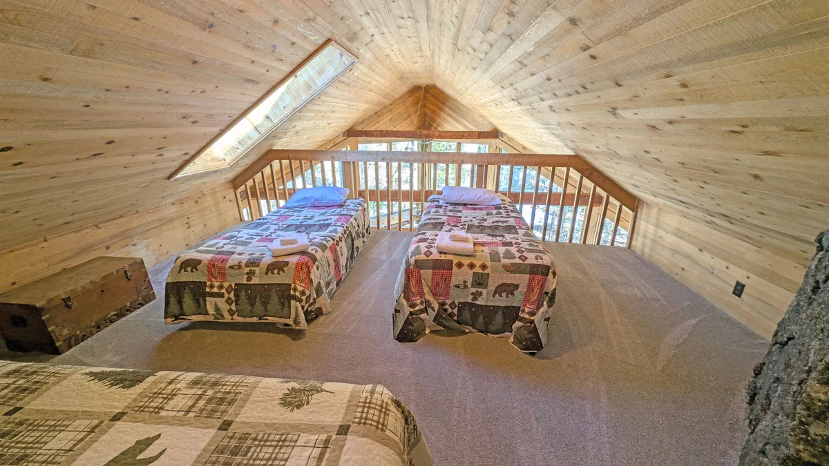 A bright, cheerful loft space featuring two twin beds with patterned quilts, wooden railings, and large windows that allow natural light to flood in. The elevated perspective and open design make it perfect for kids' slumber parties or adventurous sleepovers.