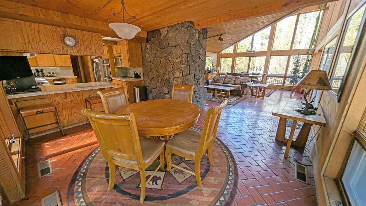 A warm, inviting dining area with a round wooden table, stone feature wall, and large windows. The natural light and cozy ambiance evoke scenes of family meals, morning coffee, or group gatherings, offering a glimpse of shared moments at the heart of the home.