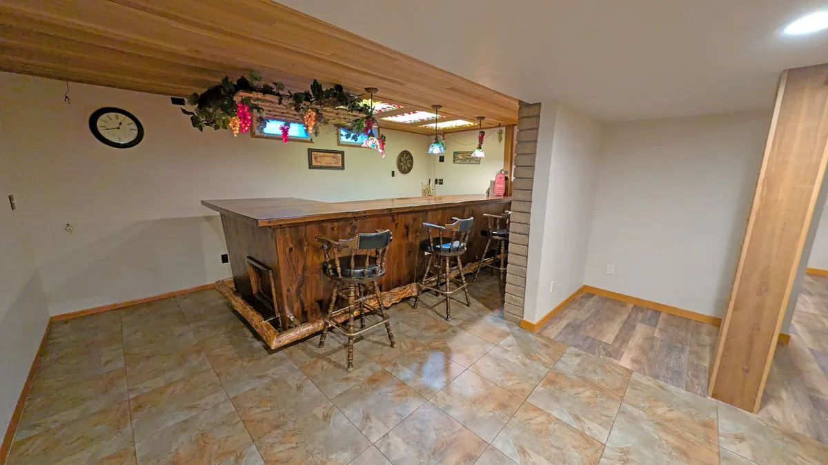 The focus here is a wooden bar with high stools, illuminated by warm decorative lighting and accented by a wall clock. This inviting space is perfect for mixing drinks, sharing laughs, and unwinding after a day of activities. The casual layout encourages conversation and relaxation.
