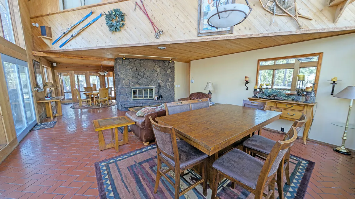 This image features a large stone fireplace as the centerpiece, surrounded by a comfortable seating area and a dining table. The room is brightened by decorative wall pieces and large windows that allow ample natural light to enter, emphasizing the warm, welcoming atmosphere.