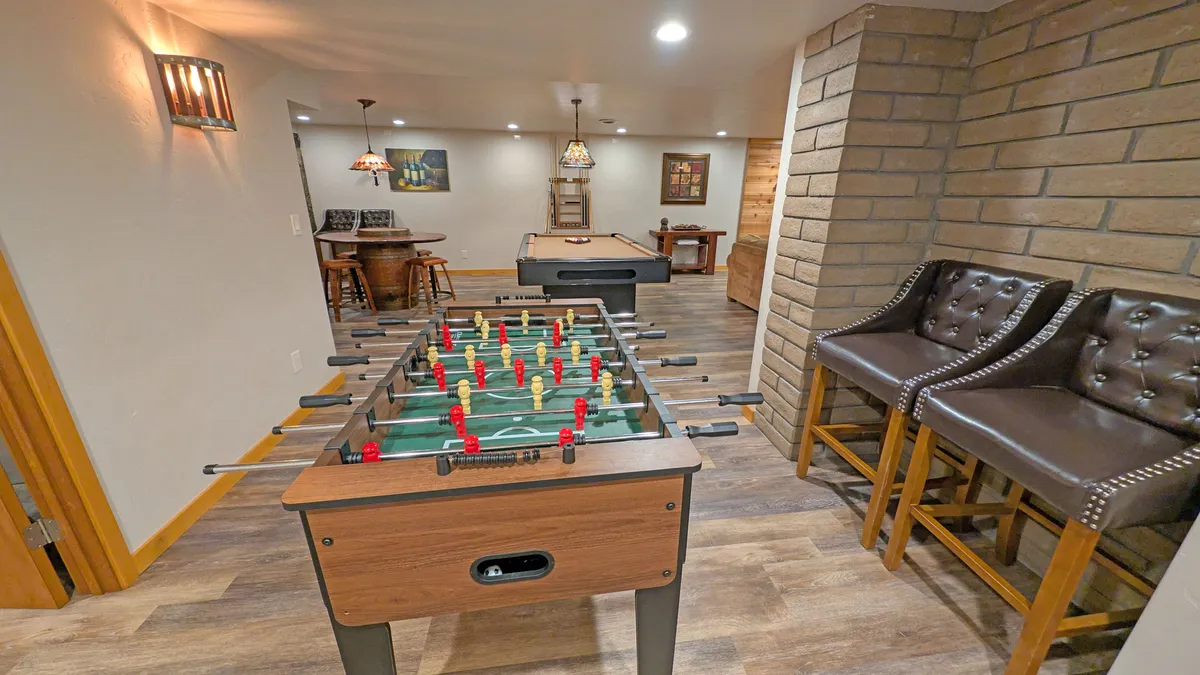 A lively basement recreation space featuring both foosball and pool tables, bar stools, and fun artwork on the walls. The layout suggests an inviting area for friendly competitions and group entertainment, perfect for families looking to bond indoors after a day outside.