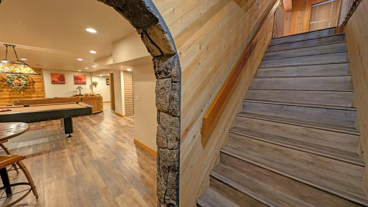 This image features a staircase leading into a cozy basement lounge with a pool table, decorative lighting, and warm wooden textures. The scene suggests an inviting environment for group activities, socializing, and game nights.