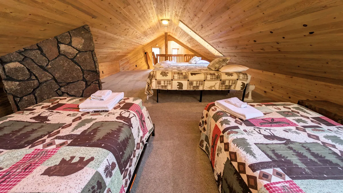 A welcoming bedroom space featuring bedding with patchwork designs, a rustic stone feature wall, and exposed wooden beams and ceiling. The loft area is visible, suggesting a cozy hideaway for stargazing or family fun. The room exudes warmth and is ideal for both relaxation and gathering.